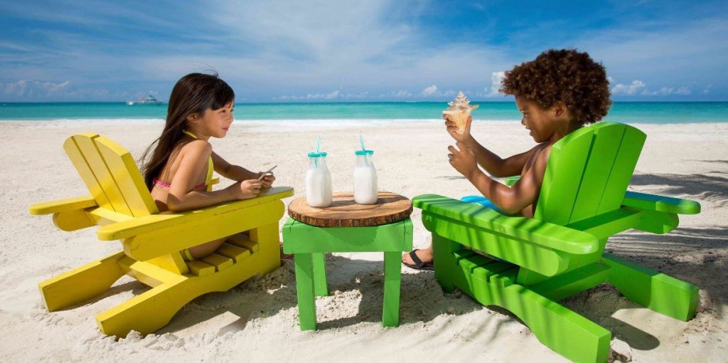 Two children sitting on the beach on vacation to the Caribbean. Both are sitting in painted wooden chairs with two bottles of milk on the table between them. Boy is holding a small conch shell