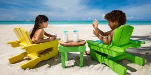 Two children sitting on the beach on vacation to the Caribbean. Both are sitting in painted wooden chairs with two bottles of milk on the table between them. Boy is holding a small conch shell